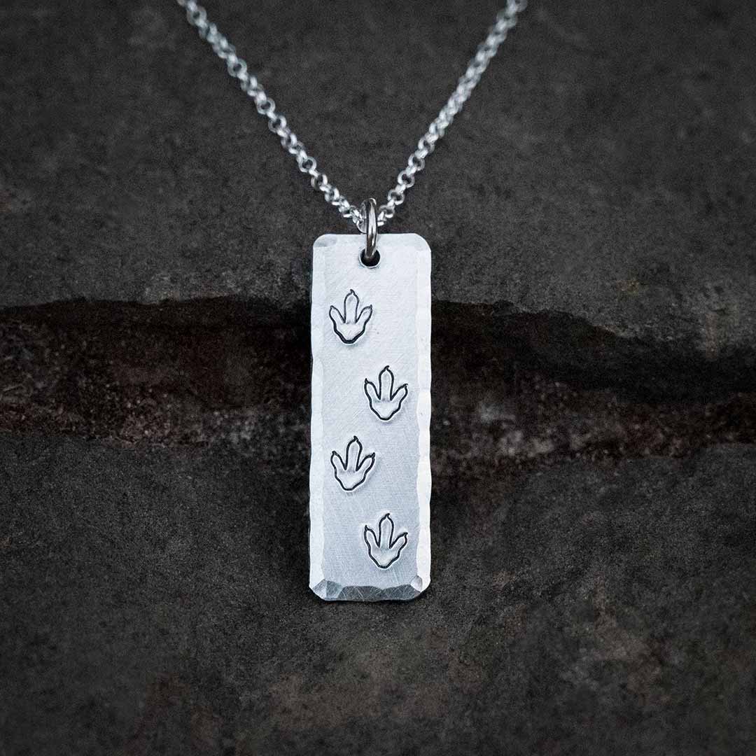 T-rex / theropod / dinosaur footprint necklace,  with the footprints hand-stamped into a vertical aluminum bar with a hammered bevel, resting on a stone background