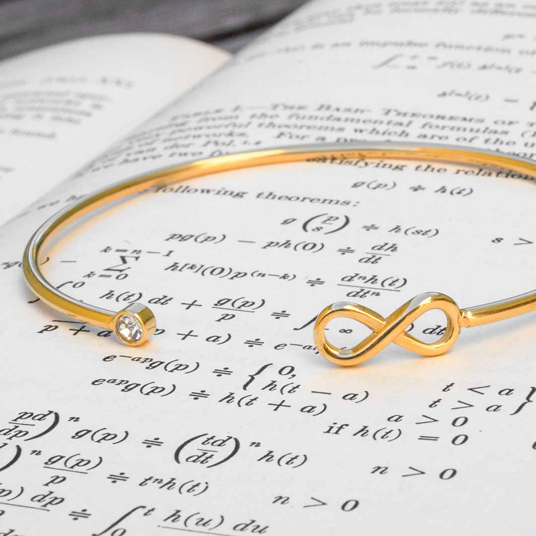 A gold-colored metal bangle bracelet featuring a small clear crystal and an infinity symbol, resting on an open book filled with mathematical formulas. Math jewelry at its finest.