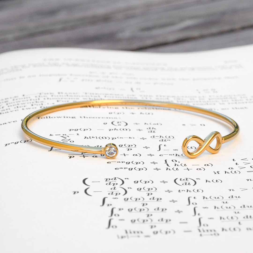 A gold-colored metal bangle bracelet featuring a small clear crystal and an infinity symbol, resting on an open book of mathematical formulas. Math jewelry at its finest.