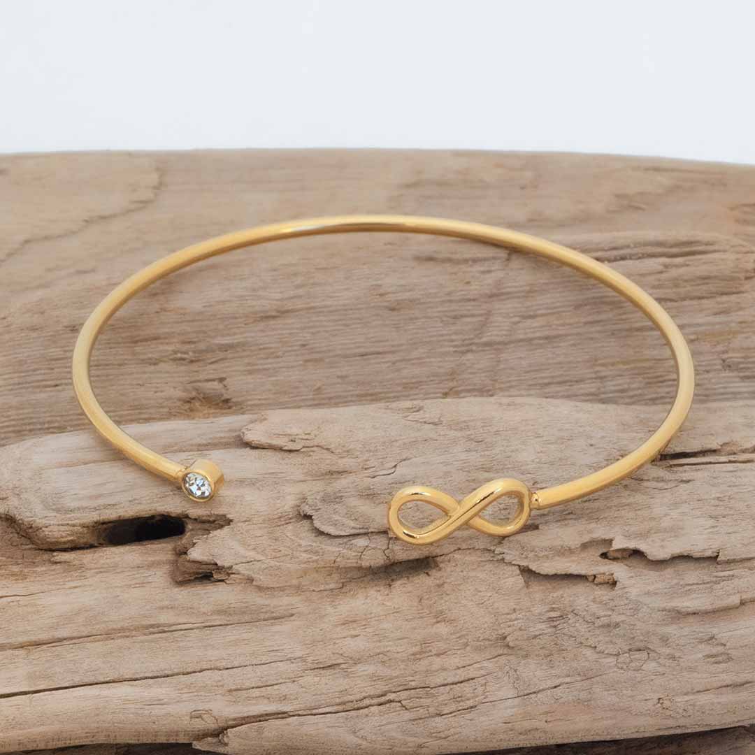 A thin gold math jewelry bangle bracelet with an infinity symbol charm and a small clear gemstone accent, displayed on a piece of weathered wood. 