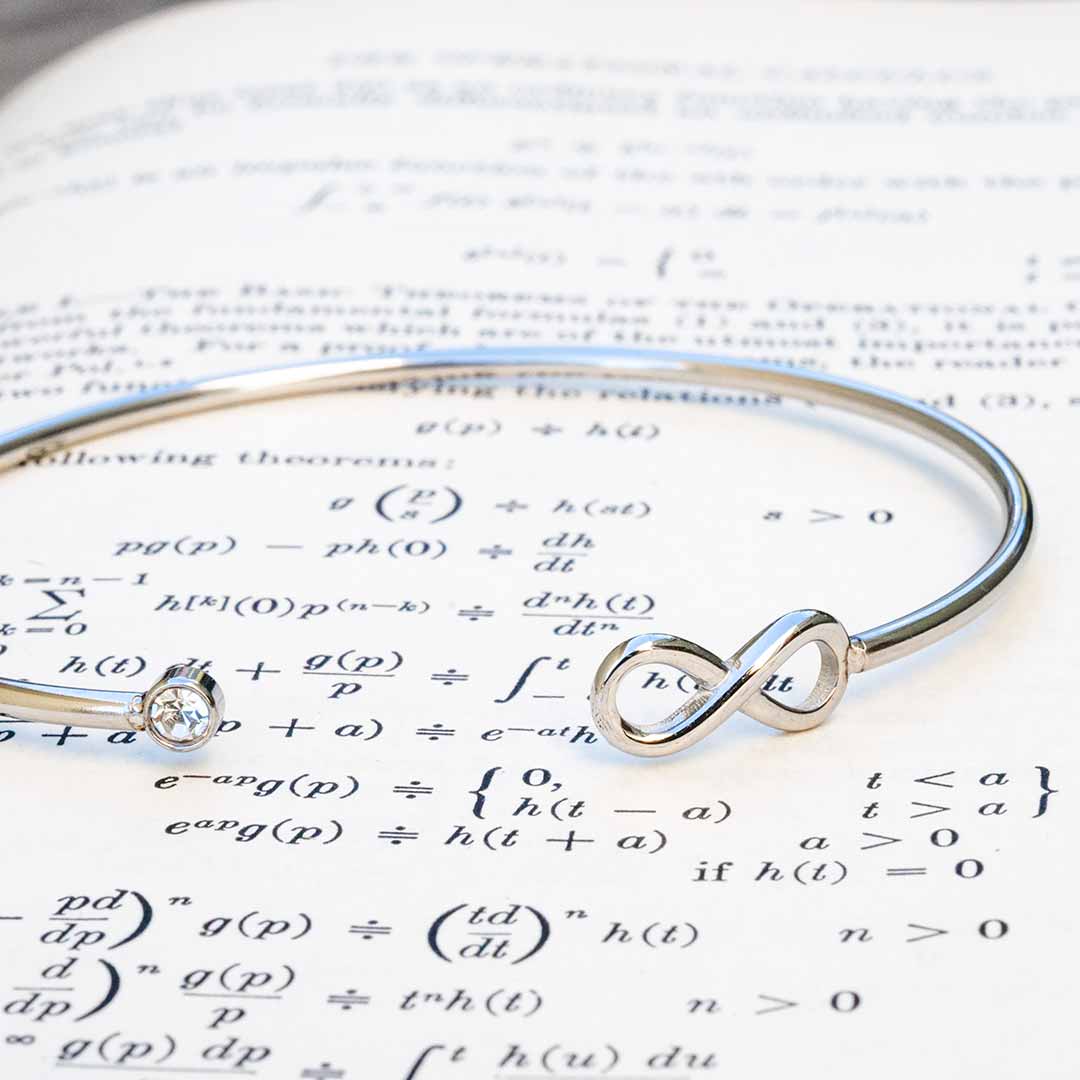 A polished silver math jewelry infinity bracelet with a single round-cut clear crystal, resting on a background of printed mathematical equations.
