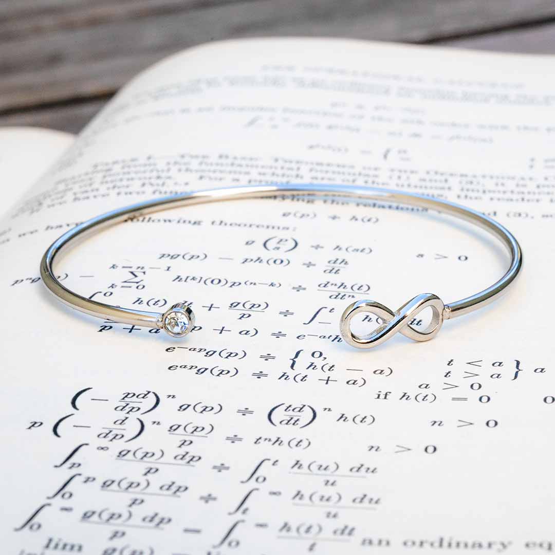 A polished silver math jewelry infinity bracelet with a small clear crystal, resting on an open book filled with mathematical equations.