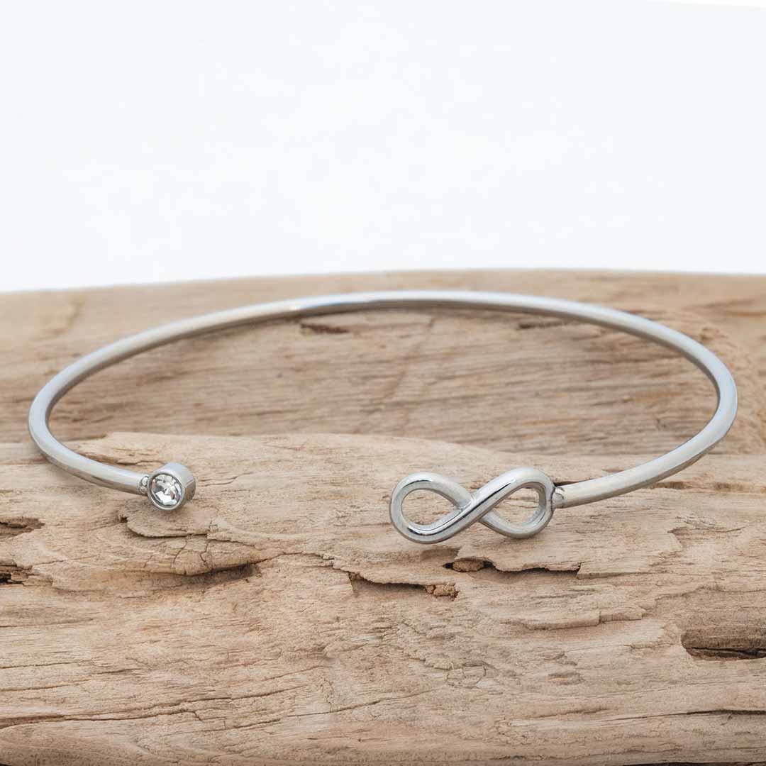 A silver-toned open cuff math jewelry bangle bracelet with an infinity symbol on one end and a small clear stone on the other, displayed on a piece of weathered wood.