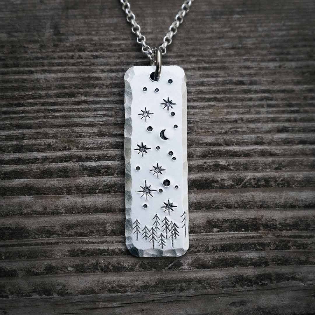 Rustic aluminum pendant hand-stamped with stars, planets, and the moon above a pine tree horizon. Necklace rests on a weathered wood plank. Beautiful astronomy jewelry for a stargazer or space science enthusiast.
