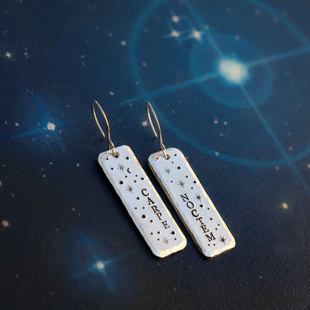 Earrings hand-stamped with the words &quot;carpe noctem&quot; and stars, resting against a background of painted blue stars on canvas.
