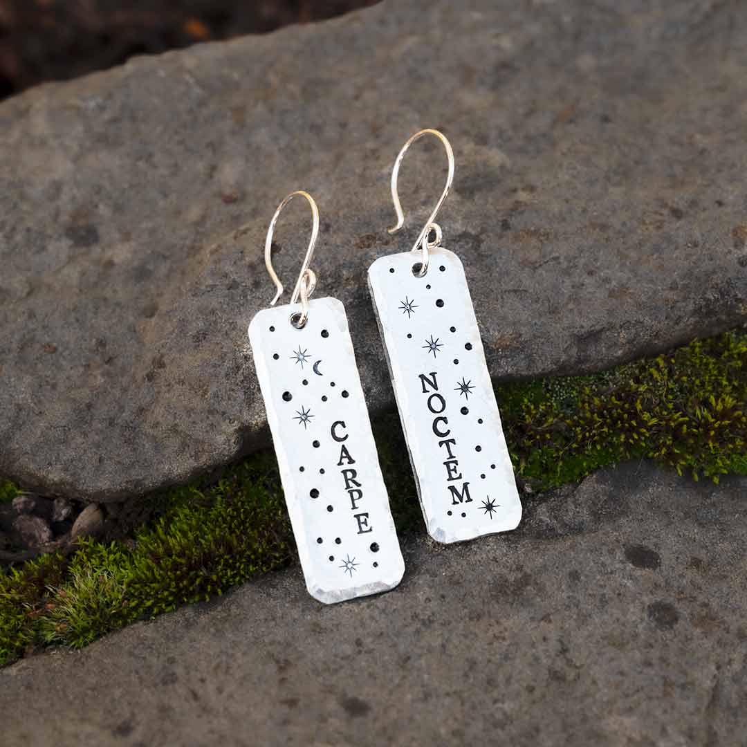 Earrings hand-stamped with the words &quot;carpe noctem&quot; and stars, resting against a background of stone and moss.