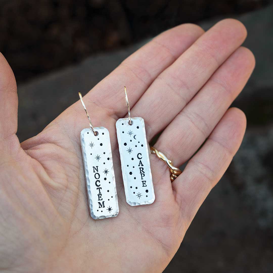 Earrings hand-stamped with the words &quot;carpe noctem&quot; and stars, being held in a hand.