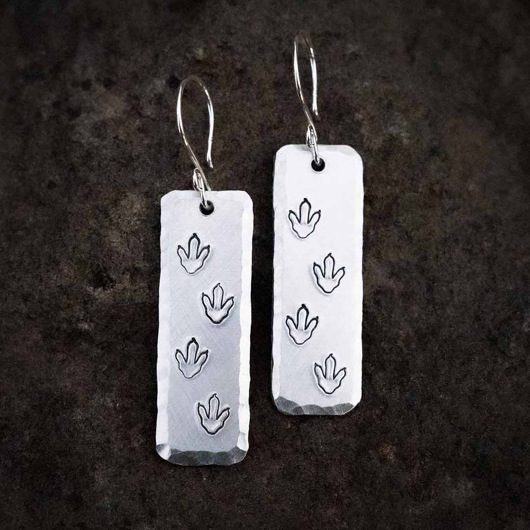 T-rex / Velociraptor / Theropod Dinosaur Footprint Earrings, made from hand-stamped metal and resting on a stone background