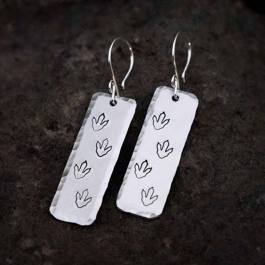 T-rex / Velociraptor / Theropod Dinosaur Footprint Earrings, made from hand-stamped metal and resting at an angle on top of a brown stone.