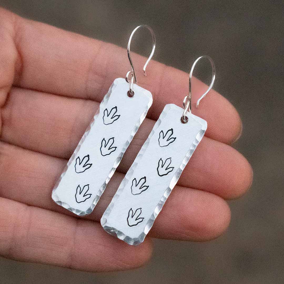 T-rex / Velociraptor / Theropod Dinosaur Footprint Earrings, made from hand-stamped metal and being held in a hand in front of a brown background.