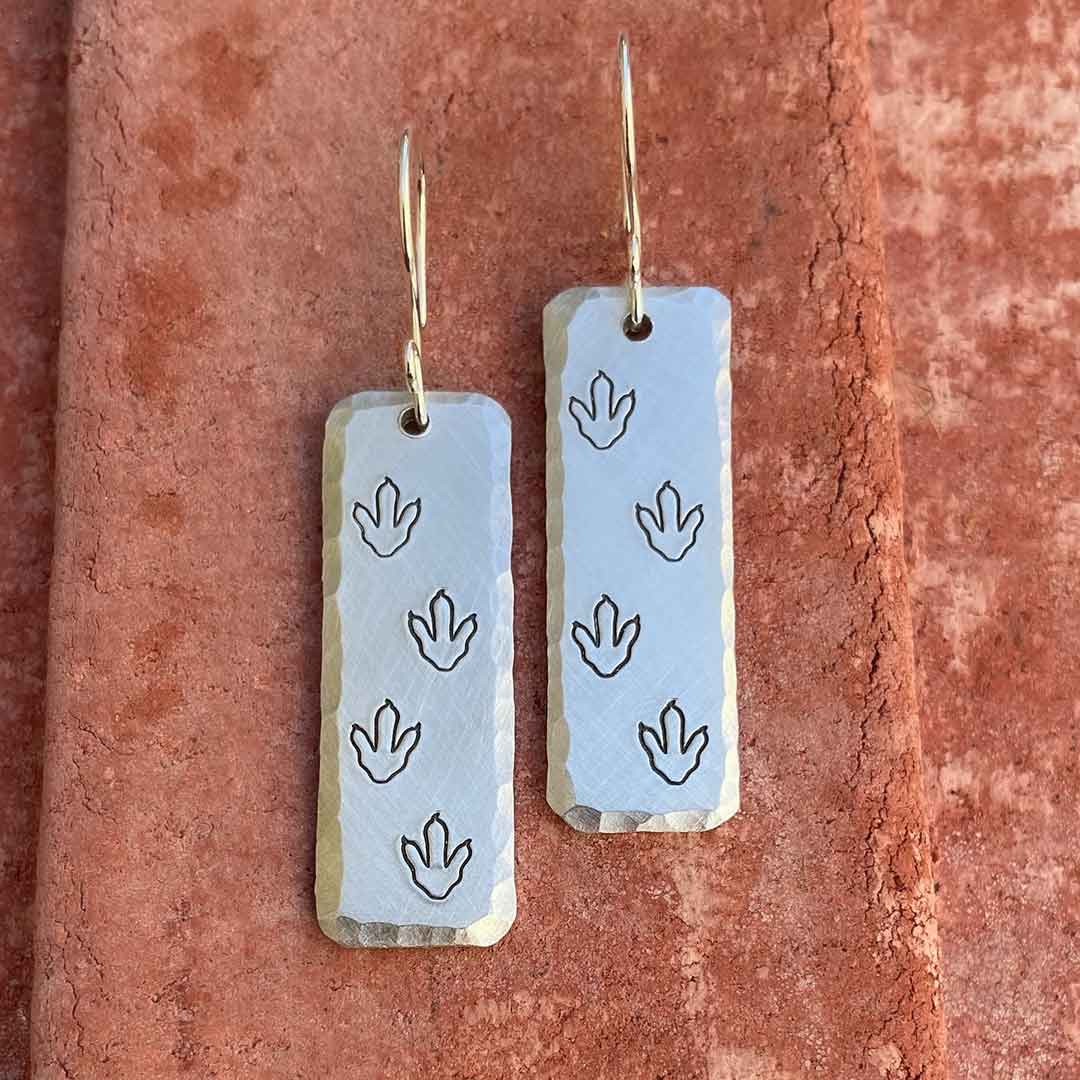 T-rex / Velociraptor / Theropod Dinosaur Footprint Earrings, made from hand-stamped metal and resting on a brick background