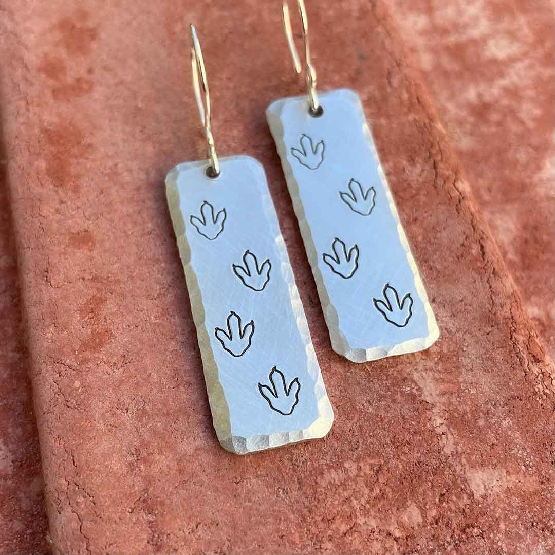 T-rex / Velociraptor / Theropod Dinosaur Footprint Earrings, made from hand-stamped metal and resting on a brick background at an angle