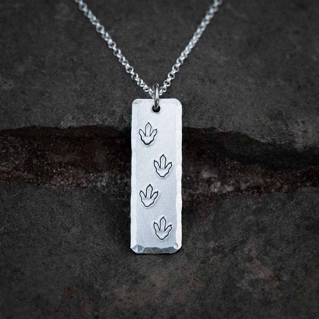 T-rex / theropod / dinosaur footprint necklace,  with the footprints hand-stamped into a vertical aluminum bar with a hammered bevel, resting on a stone background