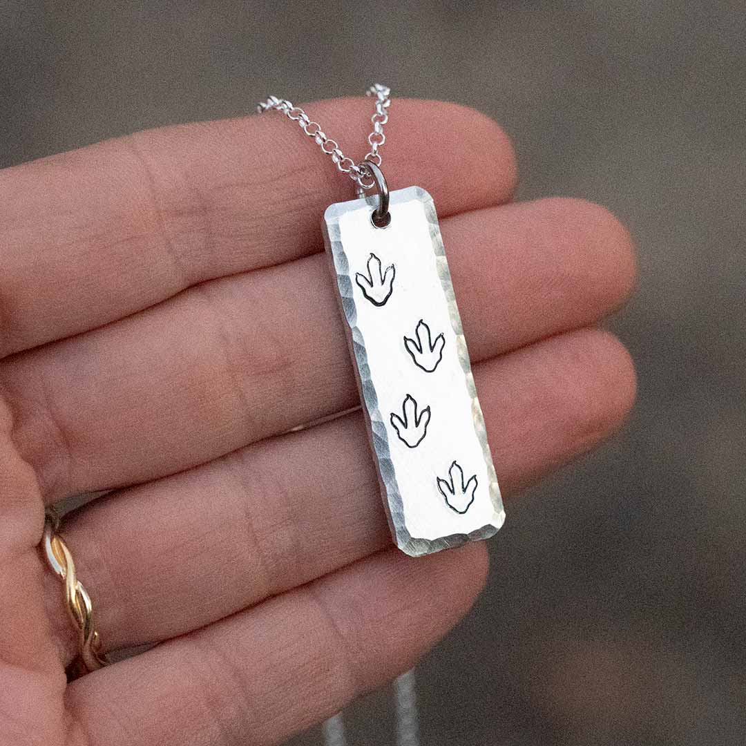 T-rex / theropod / dinosaur footprint necklace, with the footprints hand-stamped into a vertical aluminum bar with a hammered bevel, being held in a hand with a brown background.